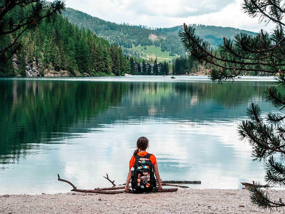 A woman sitting on a branch with a backpack, staring at the beautiful lake
