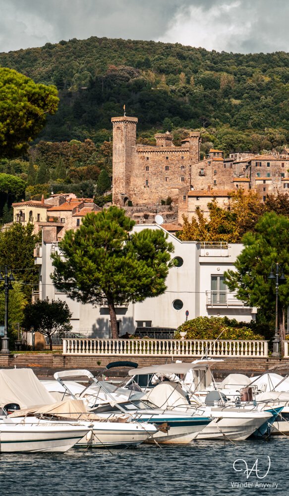 Boats docked at the Lake Bolsena harbor with a view of Rocca Monaldeschi della Cervara rising above the town, surrounded by lush green hills.